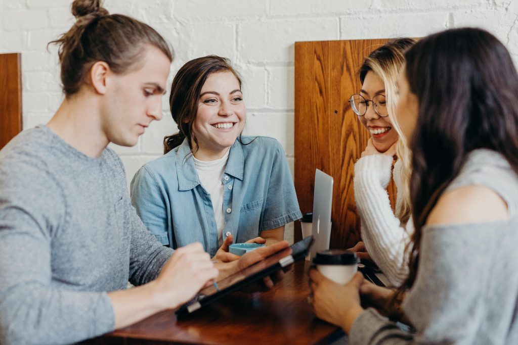 Four young people lean in around a wooden table, with two holding cups of coffee, one at a laptop, and one looking down at a tablet