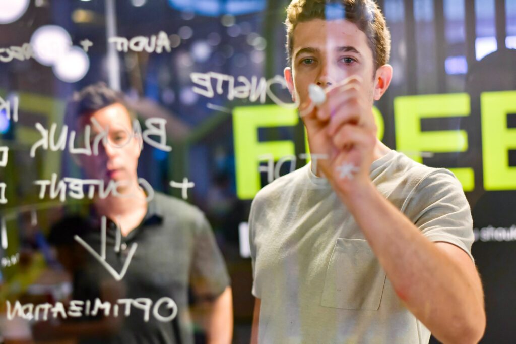 A man writes on a glass board with white texta, while another man watches over his shoulder