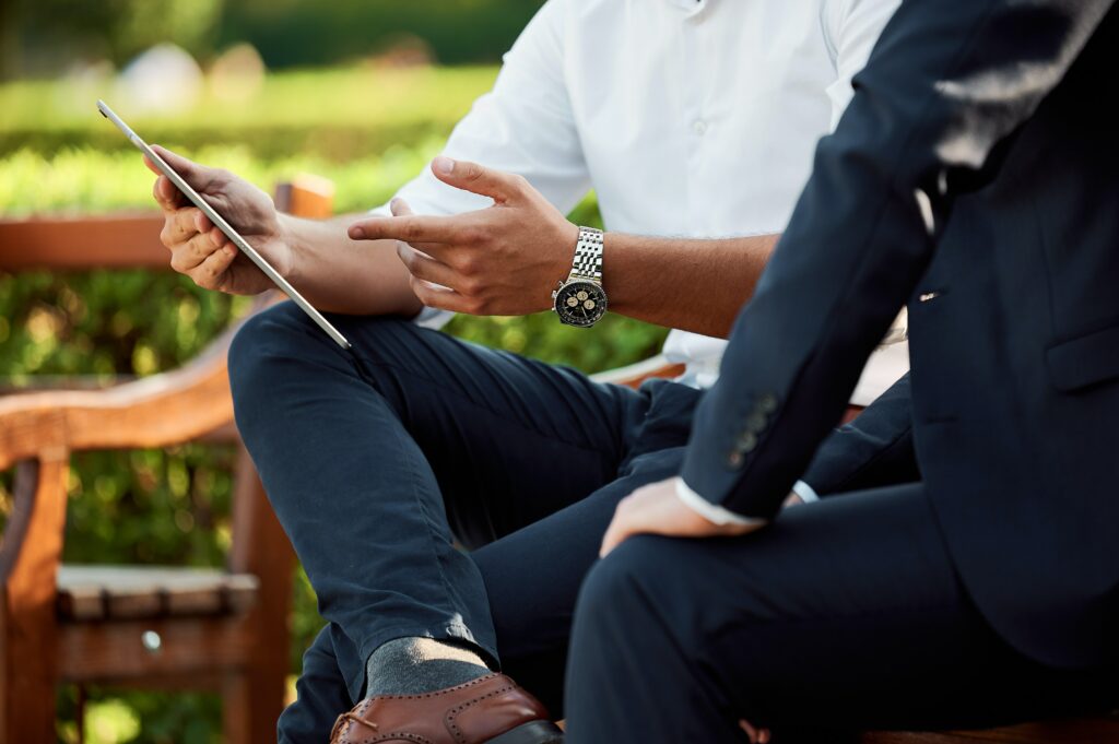 A close up shot of two men sitting on a park bench, only their torsos visible, with one of the men gesturing to a tablet