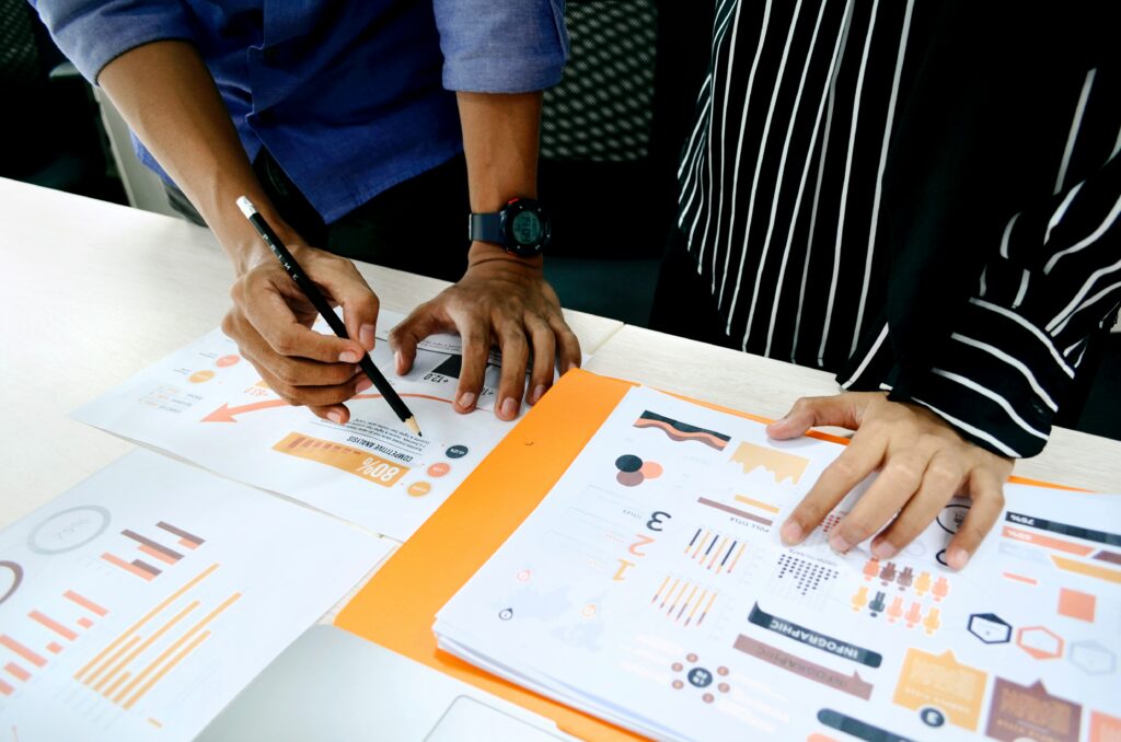 Spreadsheets strewn across a desk, with one person pointing with a pen and another person resting a hand on a document