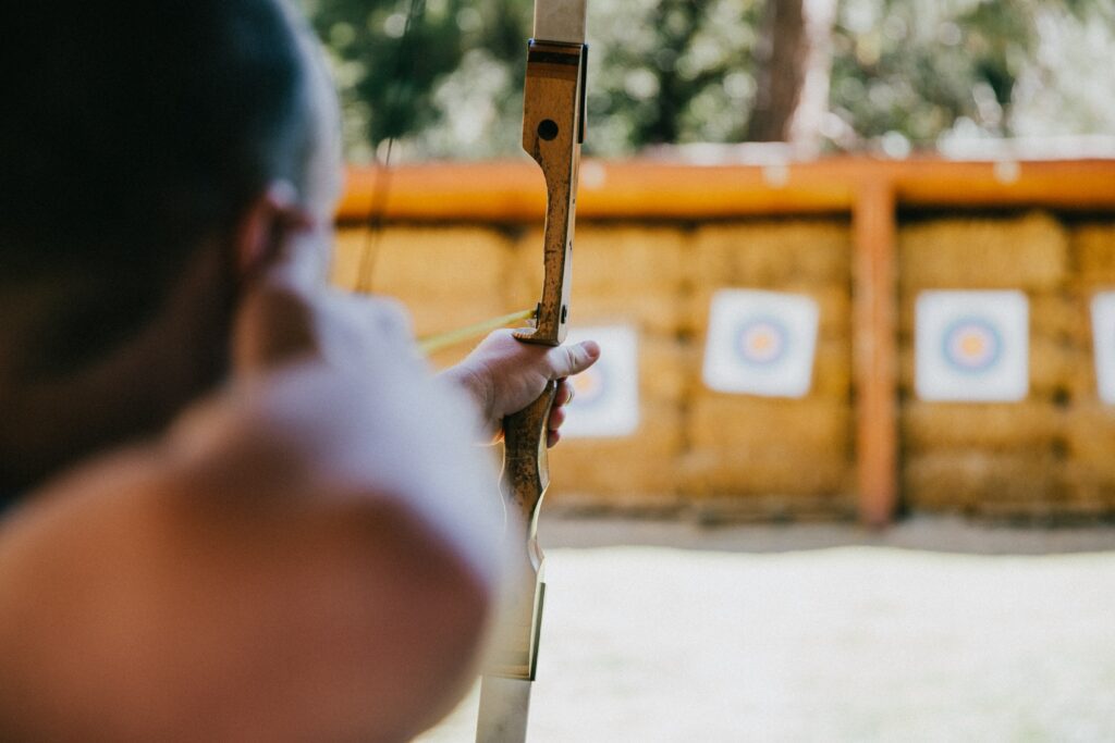 A close-up view over the shoulder of an archer as they take aim at a target pinned onto a wooden fence
