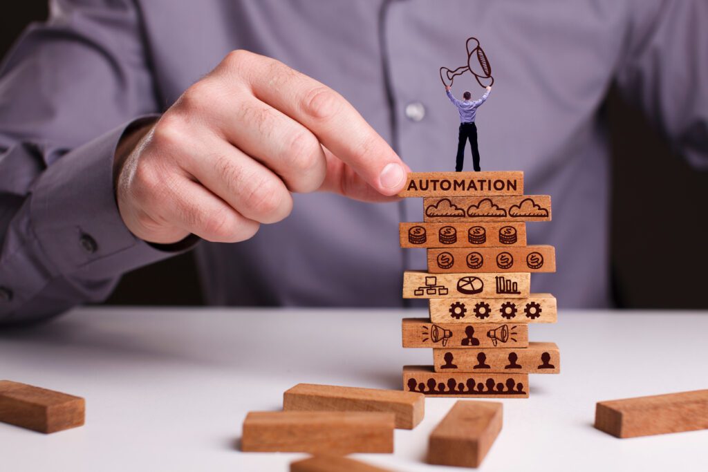 A hand selects the top block from a stack of wooden blocks, labelled AUTOMATION, on which a miniature man holding up a trophy is standing