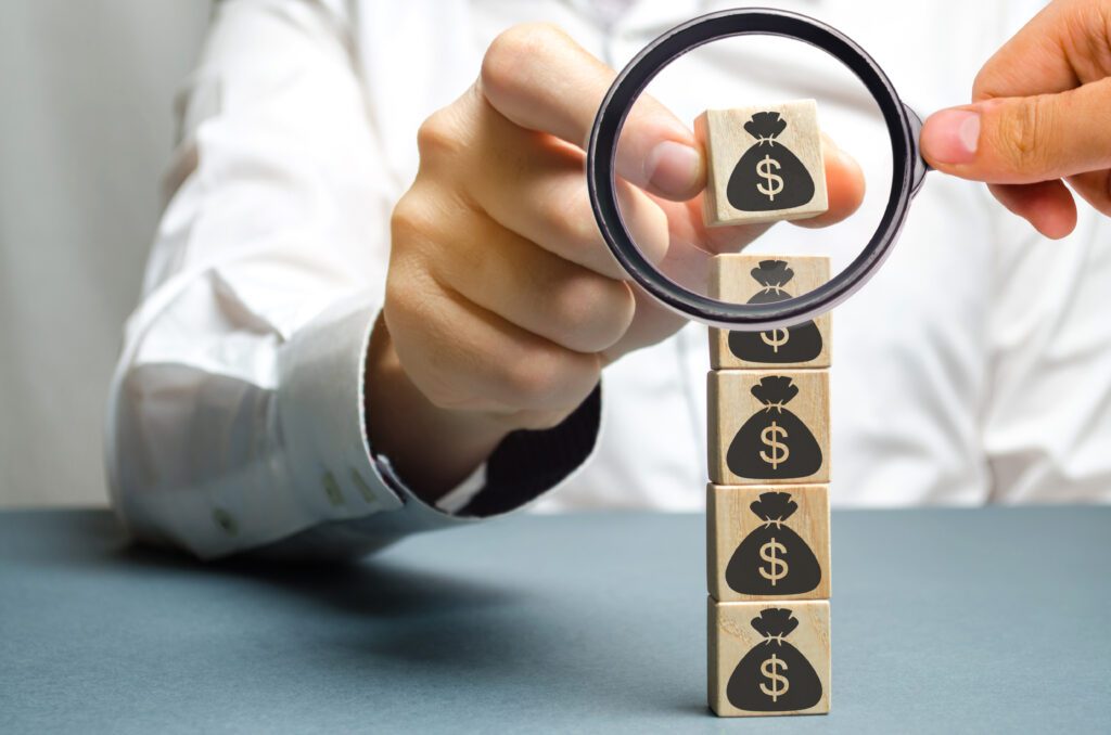 A magnifying glass is held up to the top block of a stack of wooden blocks, each with a money sack icon printed on it, while another hand picks up the block