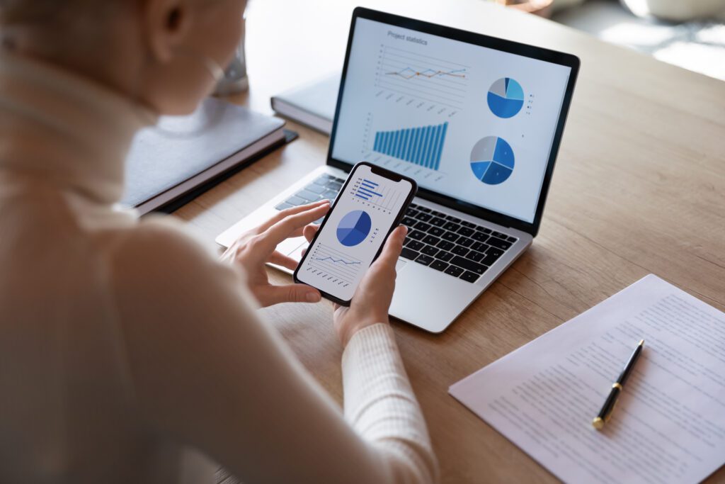 A woman sits at a laptop, holding a phone, with a range of charts on screen and a pen and document to the side