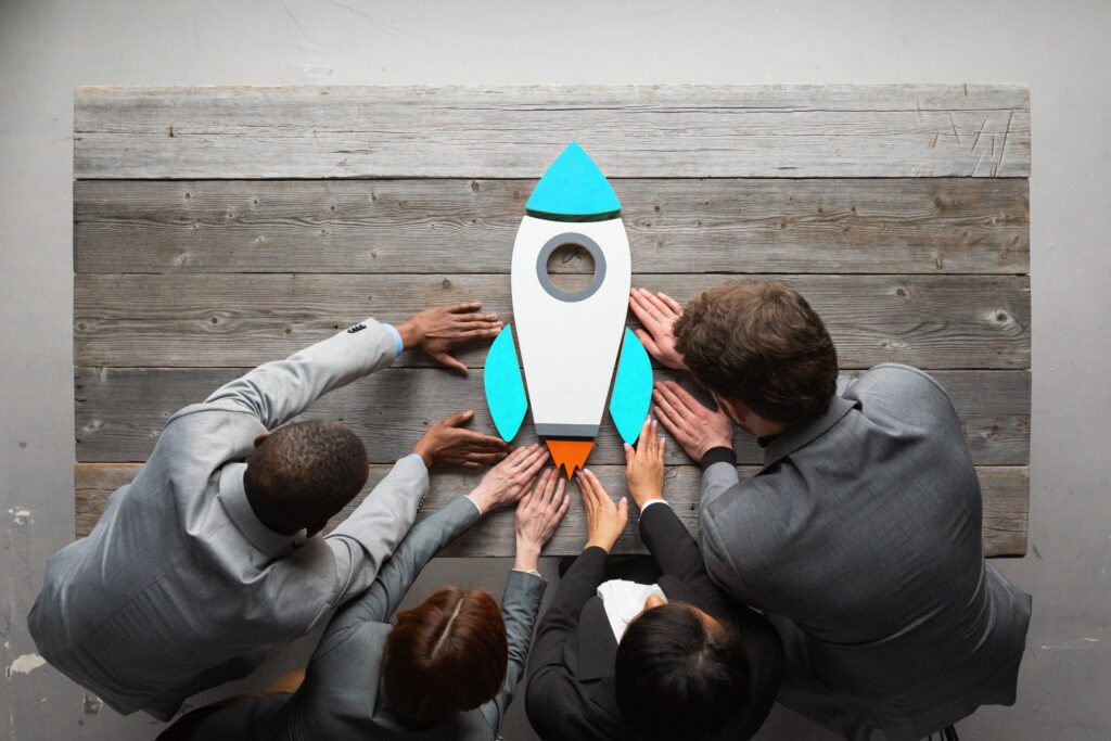 Four people in suits arrange wooden blocks into the shape of a rocket ship on a desk