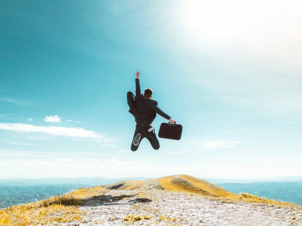 A man in a suit holding a briefcase jumps and pumps the air with his fist at the edge of a cliff, facing out to an open skyline