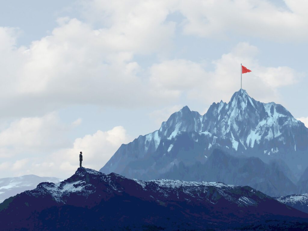 A man stands on a tall mountain, gazing at a red flag on the top of a taller mountain in the distance