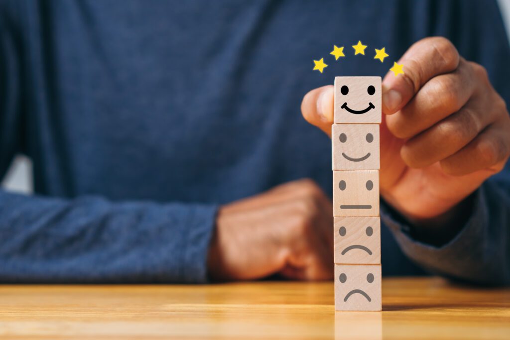 A hand selects the top block from a stack of wooden blocks, each with a face icon ranging from a sad face at the base to a smiley face at the top