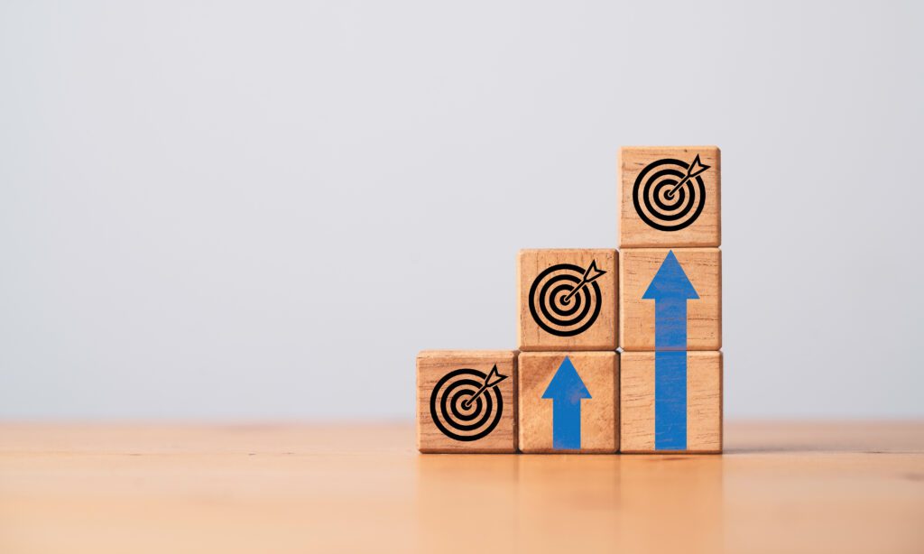 Wooden blocks stacked in an ascending staircase arrangement with target icons printed onto each top block and two upwards-pointing blue arrows
