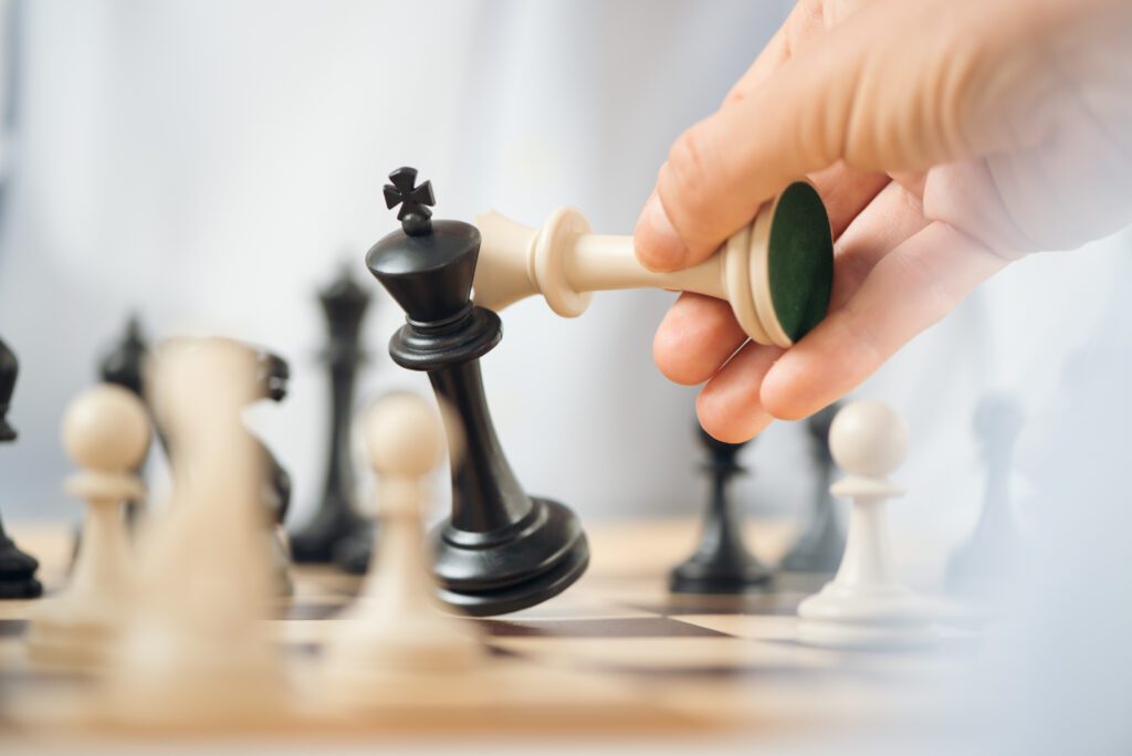 A hand uses a white chess piece to knock over a black King chess piece on a chess board