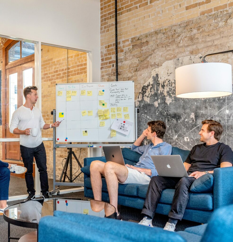 A man stands next to a whiteboard, holding a mug, and addresses two men sitting on a couch balancing laptops on their knees, both smiling