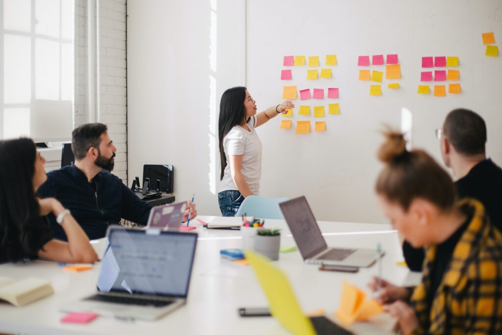 A woman points to a group of sticky notes stuck to a wall in front of four people taking notes at a desk